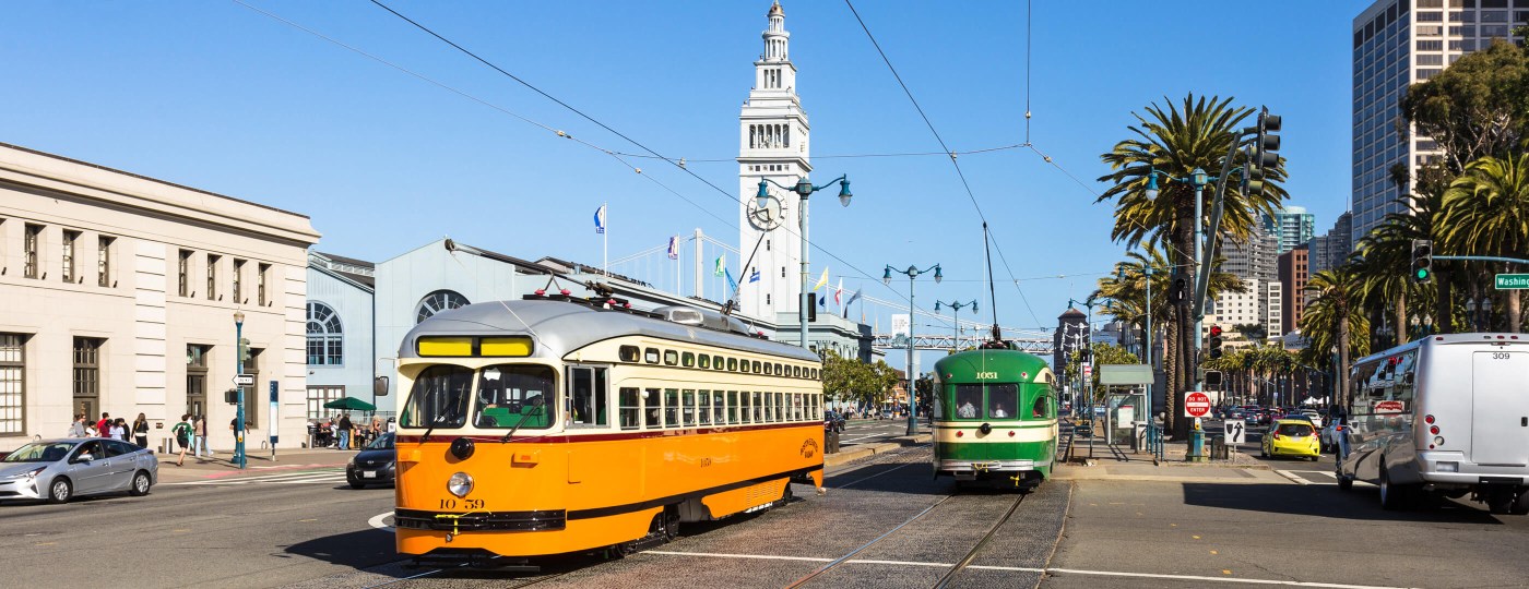 Two colorful vintage trolley cars are seen on a bustling city street in San Francisco, California, surrounded by buildings and trees with traffic and urban scenery surrounding it.