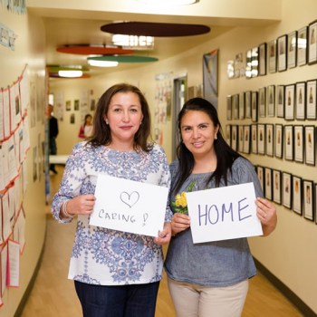 2 women in a hallway smiling with oversized cards reading: caring and home