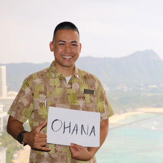 Man with a colorful shirt with Waikiki Beach behind him, holding an oversized card reading: ohana