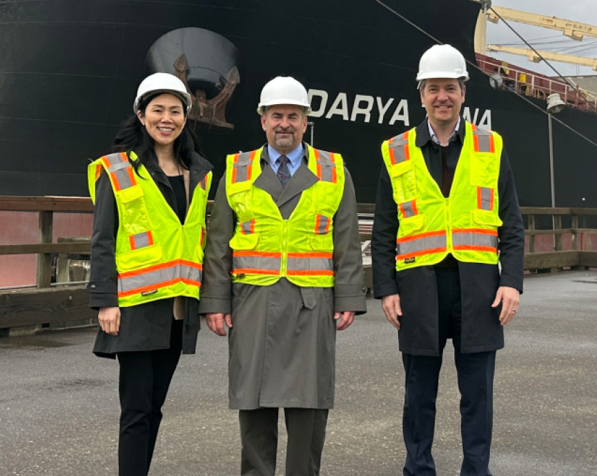 three executives in hard hats and safety vests with business attire underneath posing for a picture in a harbor in Oregon with a shipping vessel in the background