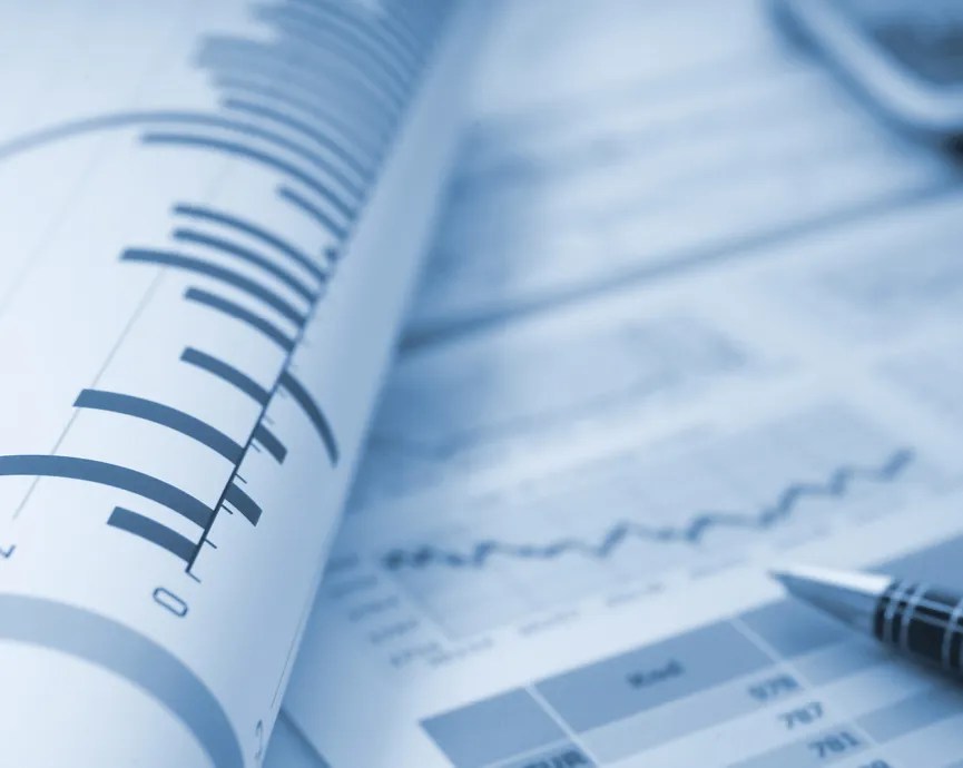 two pages of indistinct spreadsheets with various graphs and charts and the tip of a pen in the bottom right-hand corner