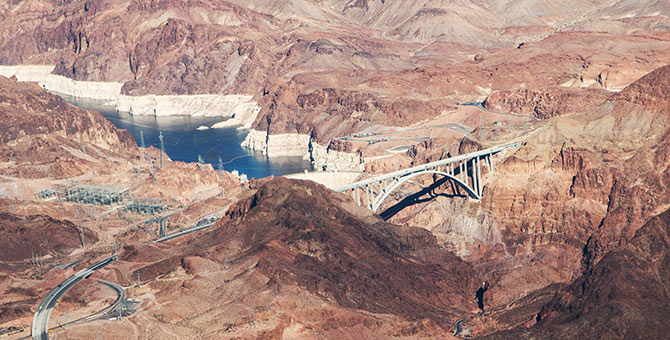 Aerial view of Hoover Dam, showing a low water level as well as the surrounding craggy landscape, and the bridge.