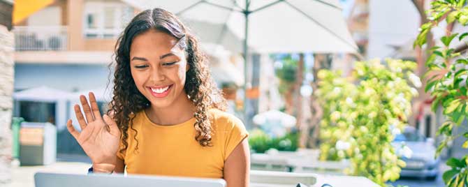 A woman outside beneath an umbrella waving at her computer