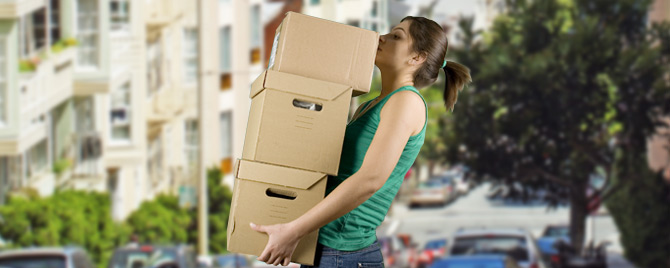 Young woman carrying several large moving boxes across the street.