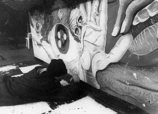 Brian laying on the floor as he works on a mural that includes hands, an eye, and a skull